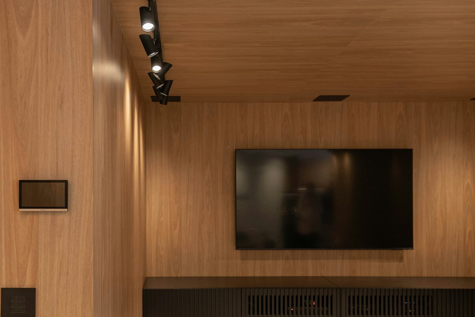 A wood-panelled media room with a wall-mounted touch panel and keypad, ceiling track lights, and a large display above an equipment credenza.