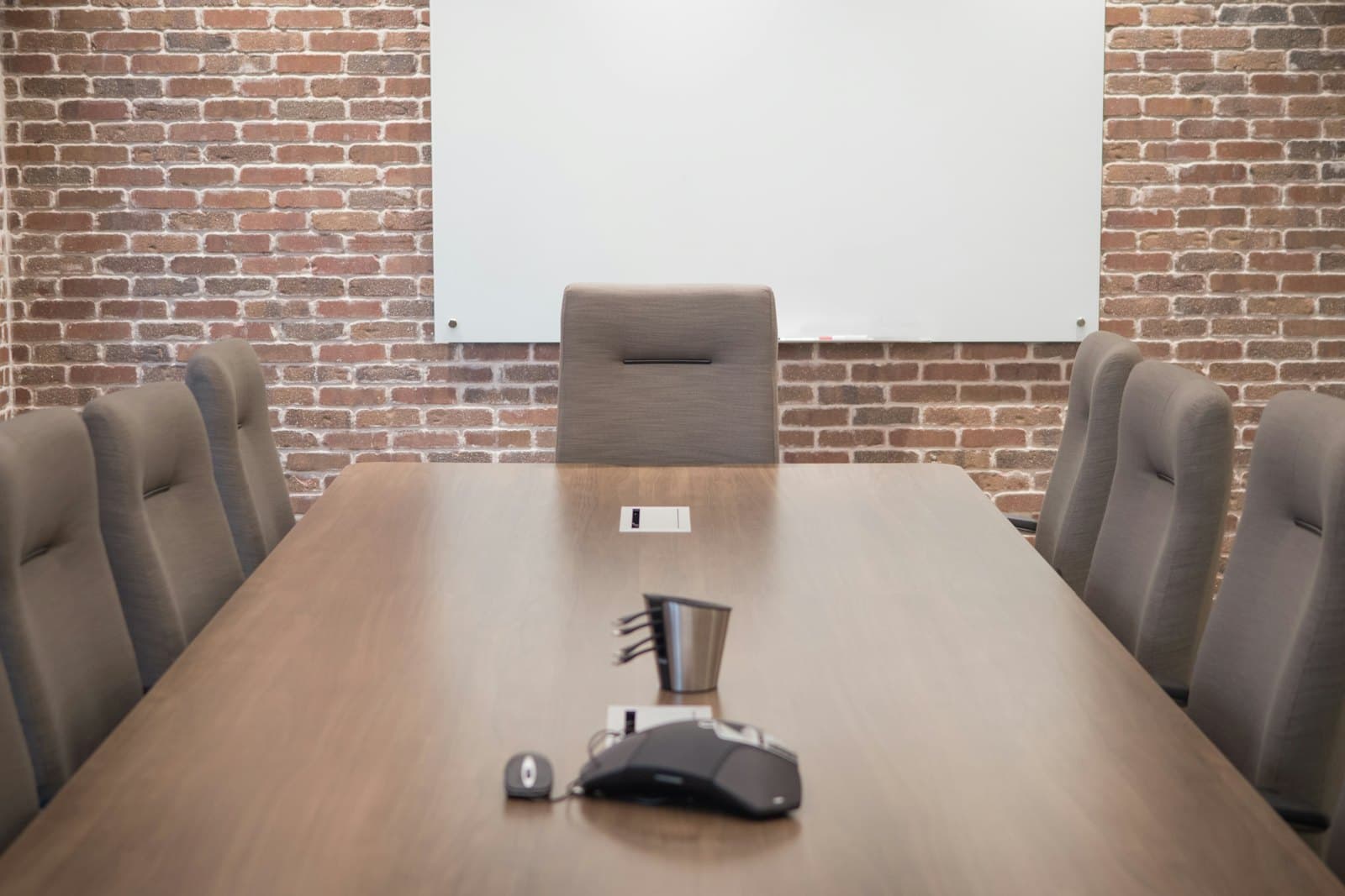 An empty conference room: a long wooden table with a conference phone and cable cubby, chairs on both sides, and a wall-mounted screen against brick.