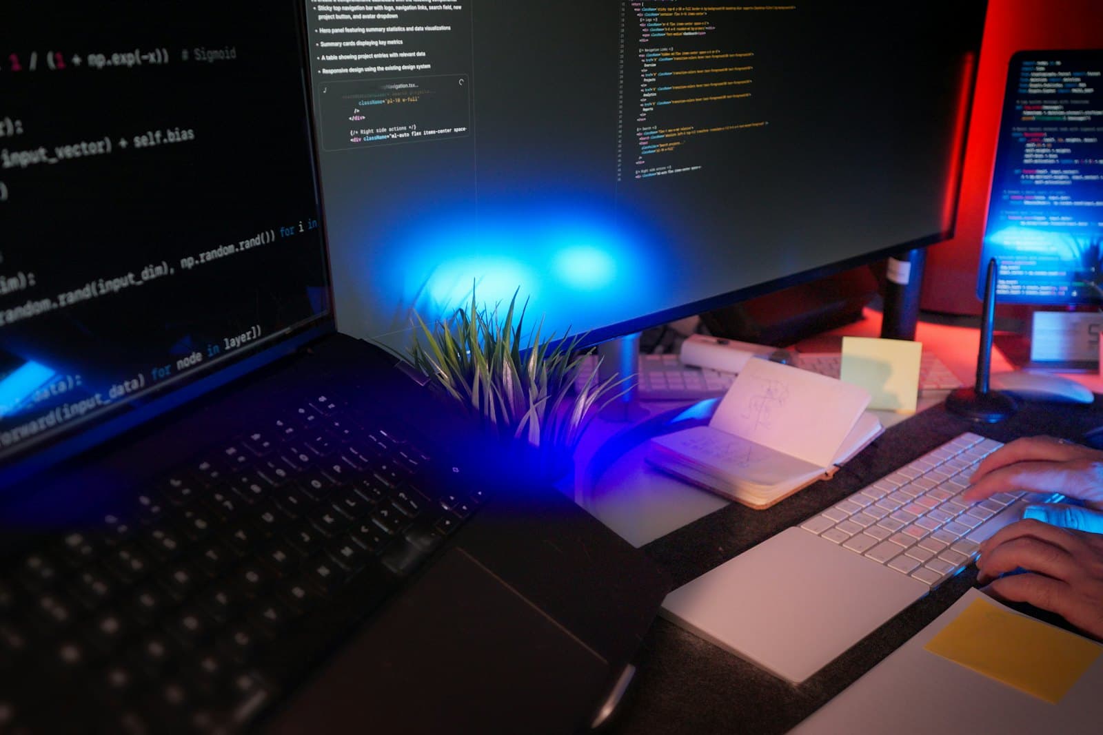 A programmer's hands on a keyboard at a desk with several monitors showing code, a laptop and an open sketchbook.